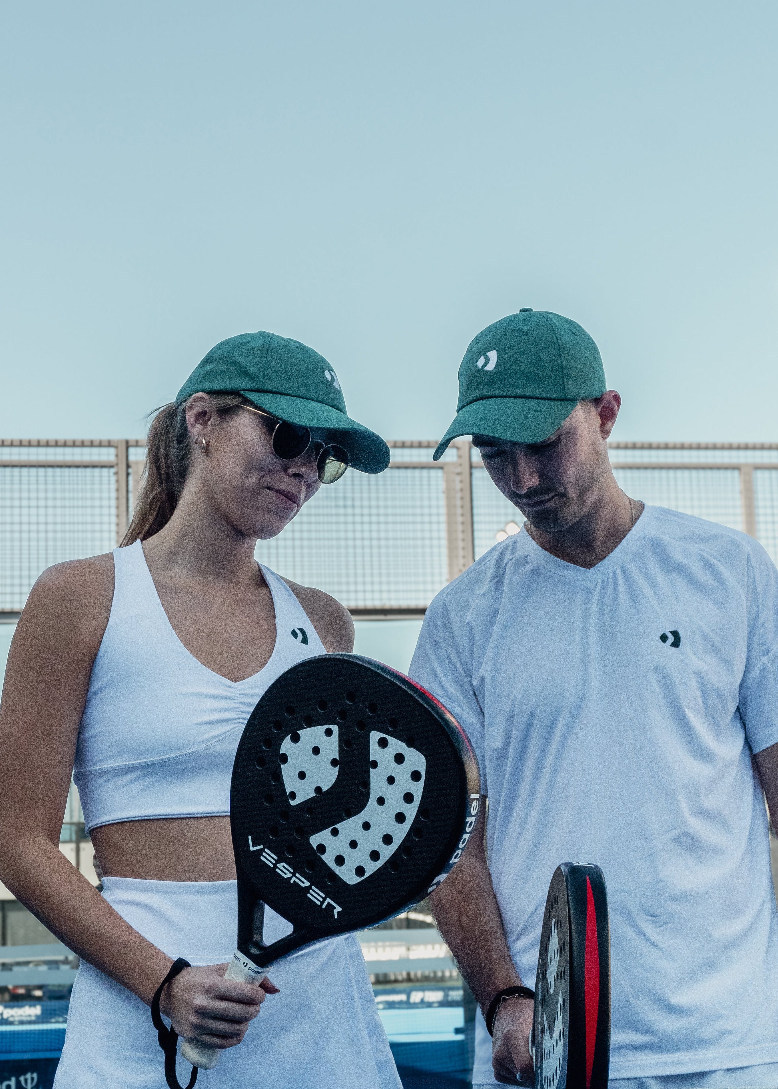 Two people on a padel tennis court holding paddle sports equipment. The padel racket is being held on a padel court. paddle players in the sun. They are wearing padel clothing and padle racquets. Two premium padel rackets are also featured.