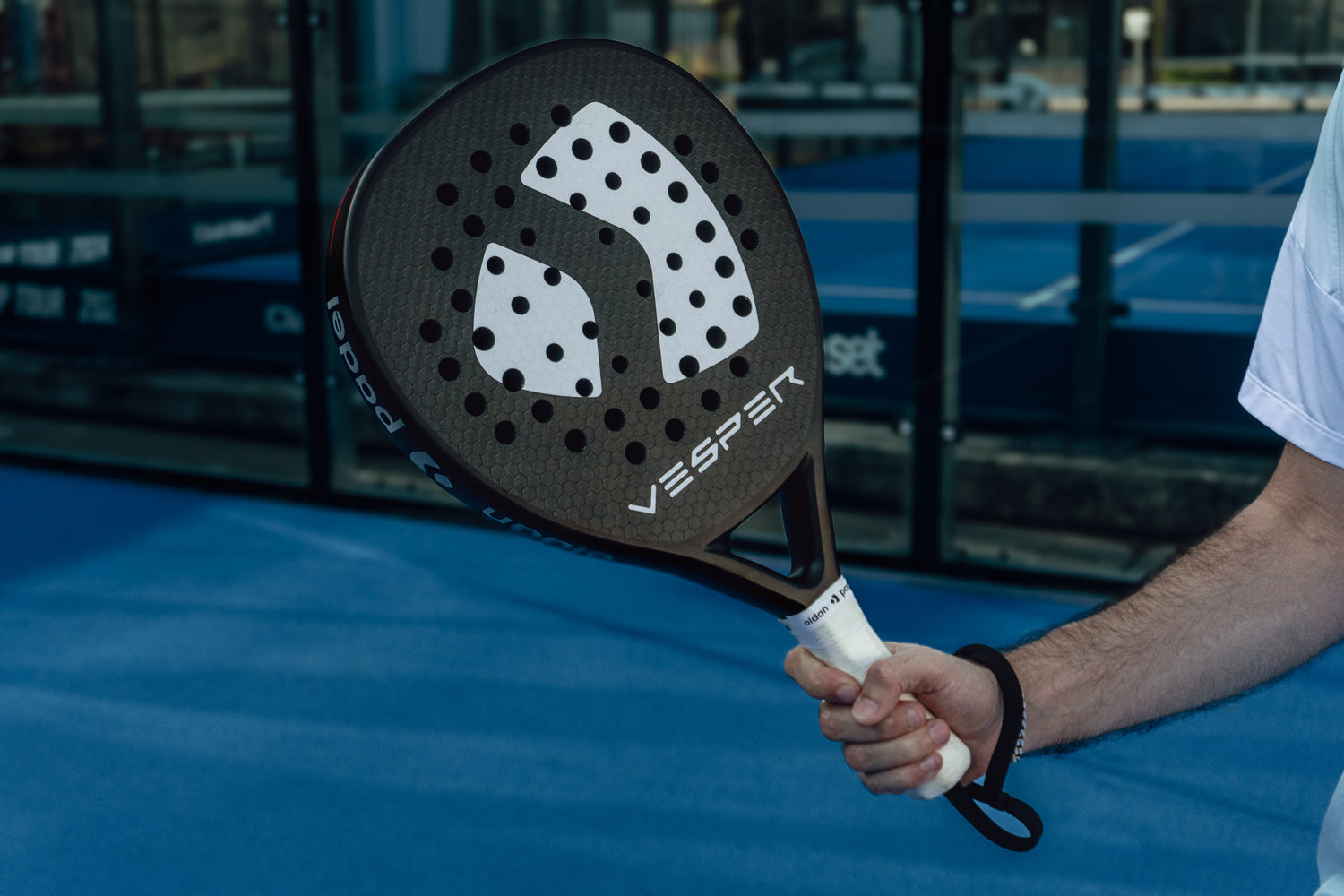 Paddle held by a person on a blue surface with 'Vesper' branding. A padel racket on a padel tennis court. 