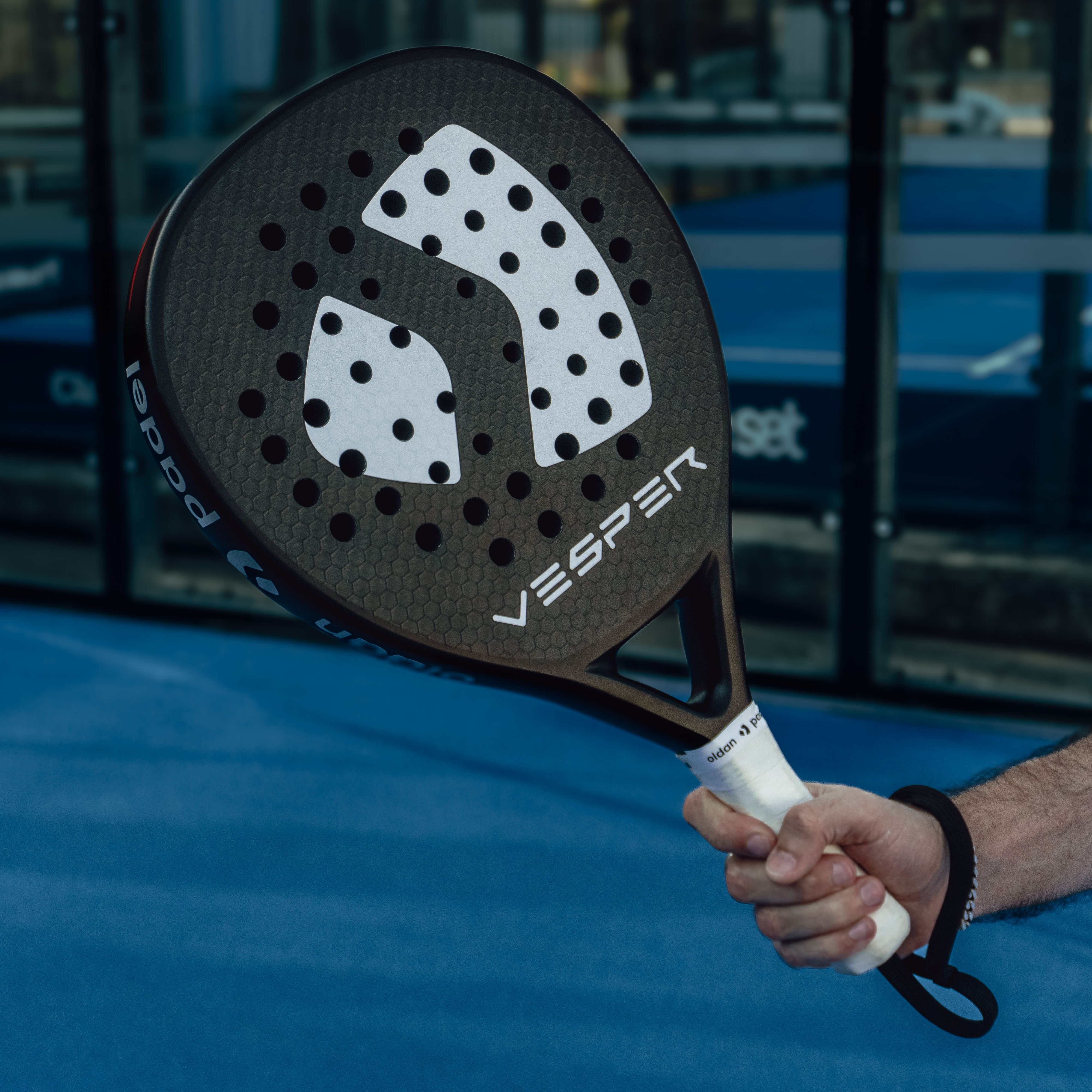 Paddle held by a person on a blue court with 'Vesper' branding. Padel racket on a padel court. Premium Padel Racket.